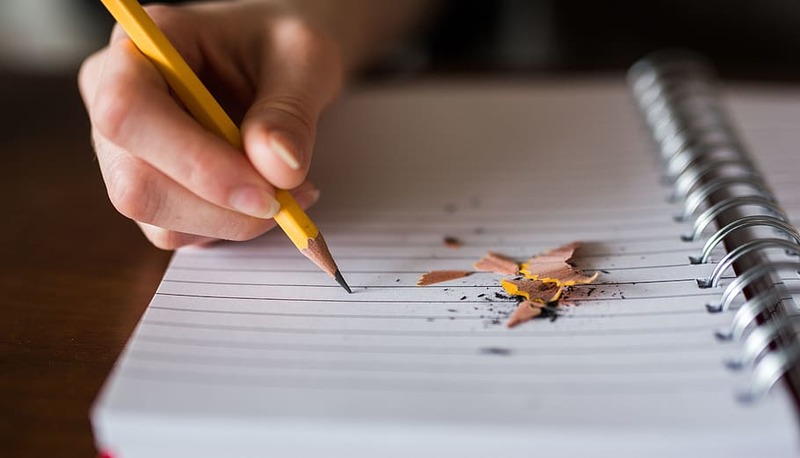 Una mano tiene una matita gialla sopra un quaderno, circondata da trucioli di legno. Focus sulla scrittura e creatività.